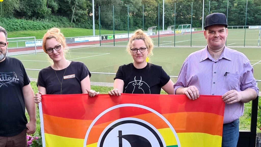 Elaine und Maria Bach, Patrick Bodden und Stefan Borggraefe halten zusammen eine Queeraten-Flagge