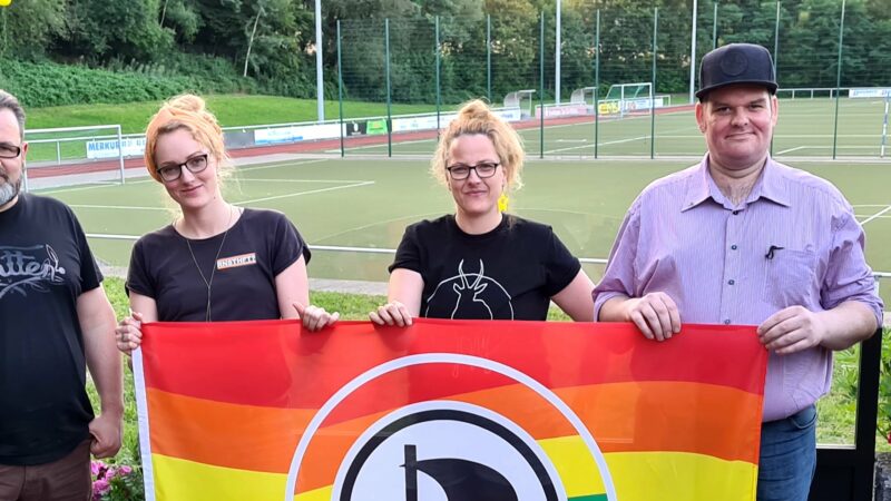 Elaine und Maria Bach, Patrick Bodden und Stefan Borggraefe halten zusammen eine Queeraten-Flagge