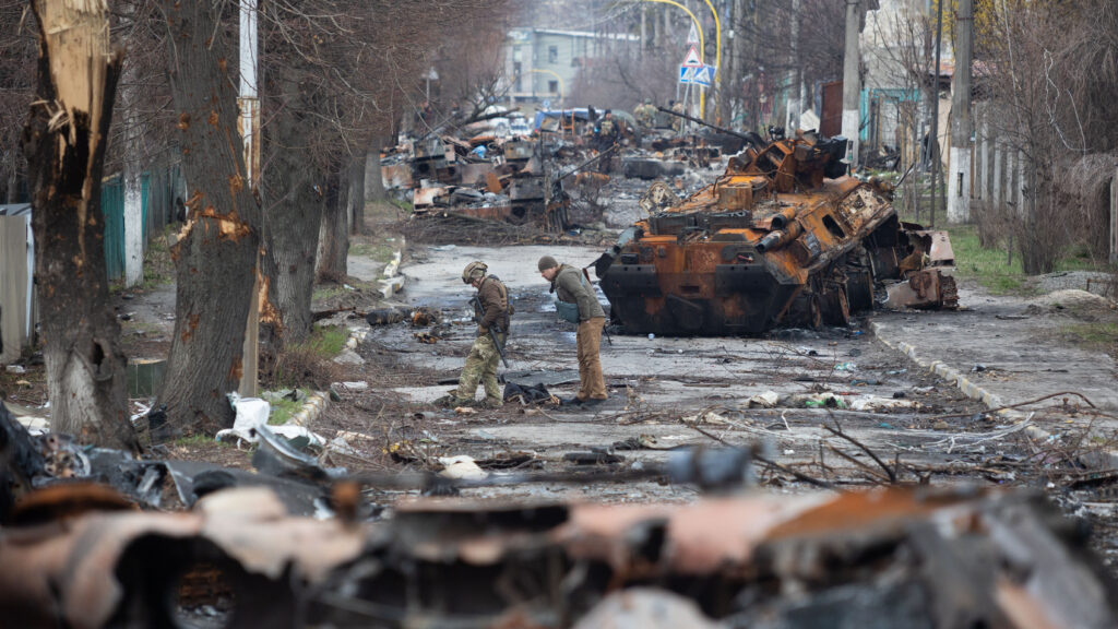 Bucha Hauptstraße nach der Russischen Invasion. Man sieht zerstörte Autos, Militärfahrzeuge und Bäume. Soldaten untersuchen die Straße.