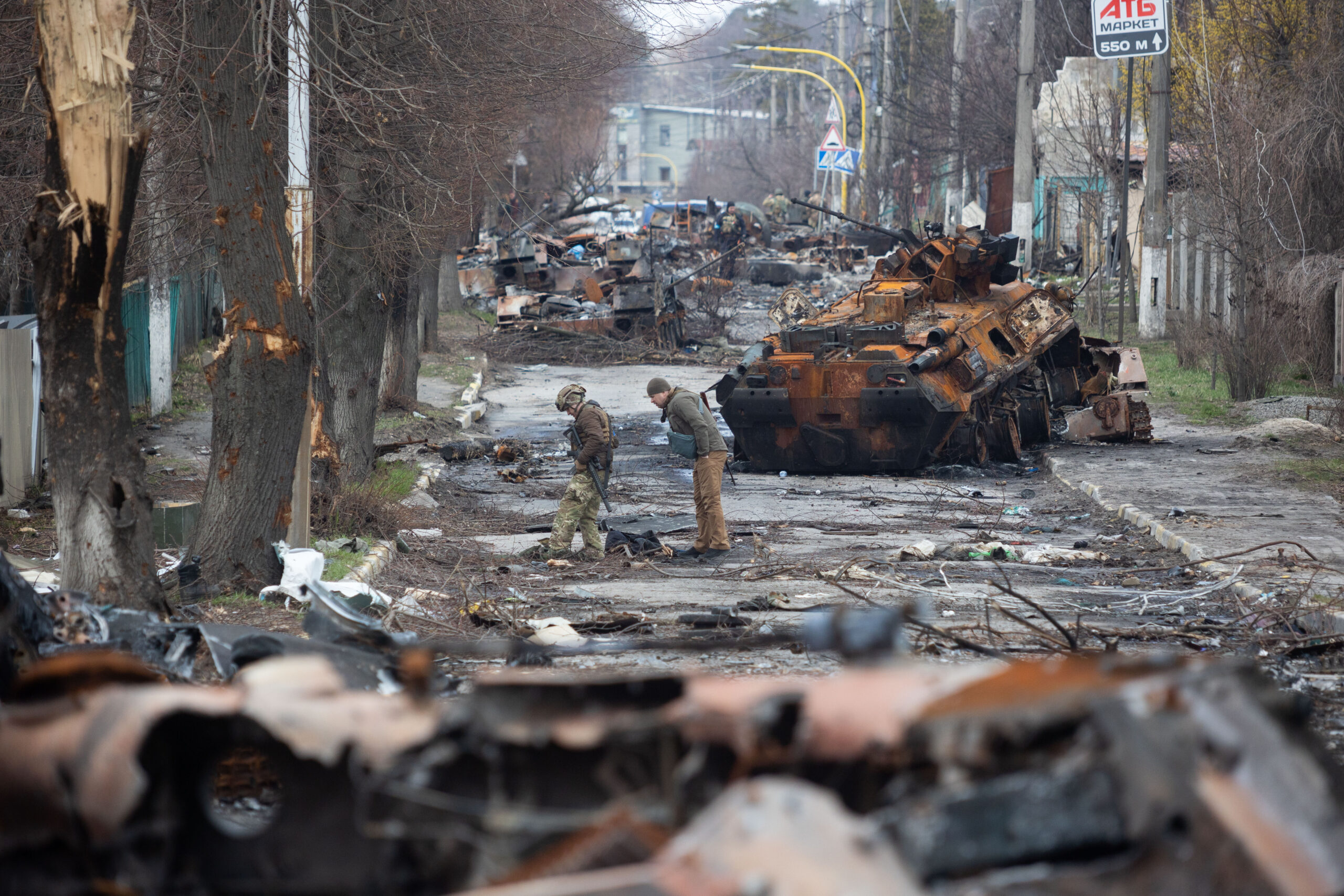 Bucha Hauptstraße nach der Russischen Invasion. Man sieht zerstörte Autos, Militärfahrzeuge und Bäume. Soldaten untersuchen die Straße.