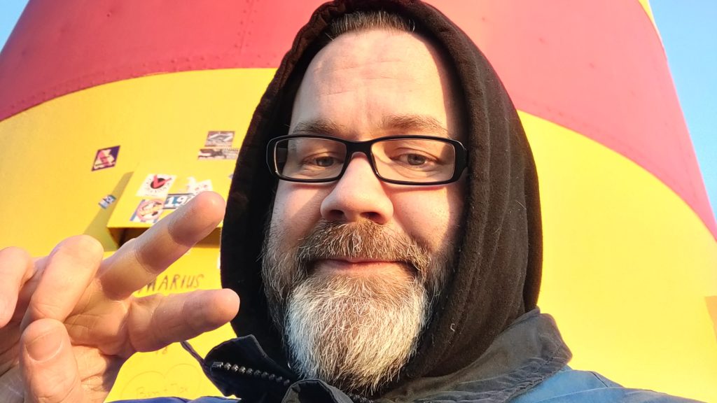 Stefan Borggraefe mit Hoodie vor einem Leuchtturm. Mit der rechten Hand macht er ein Victory-Zeichen.